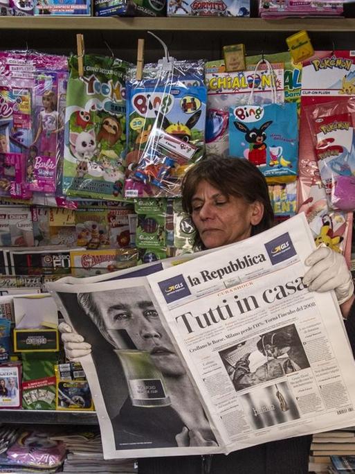 Eine Zeitungsverkäuferin in einem Kiosk in Italien hält einen Zeitungstitel hoch:"Tutti a casa" ("Jeder zu Hause") in Salerno, Italien am 10.März 2020