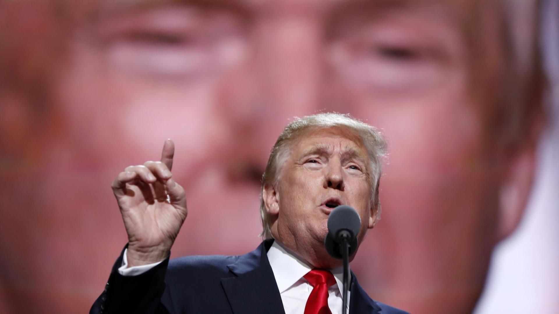 Donald Trump mit erhobenem Zeigefinger vor einem großen Bild von ihm selbst