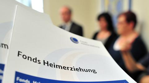Ein Prospekt mit der Aufschrift "Fonds Heimerziehung" liegt auf einem Tisch vor einer Gruppe von Menschen.