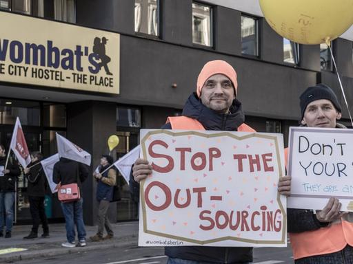Demonstranten stehen vor einem Hostel und halten Transparente mit Slogans wie "Stop the Outsourcing" hoch.