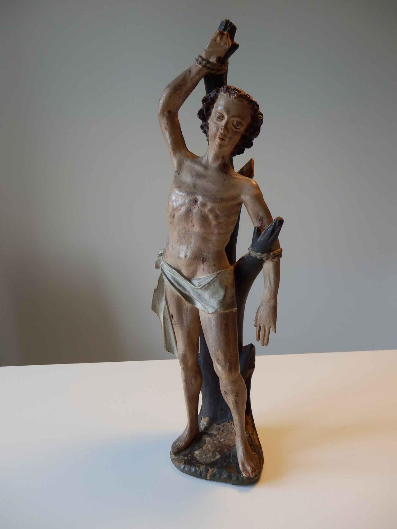 Holzskulptur des Heiligen Sebastian, der nur einen Lendenschurz trägt, an einen Baum gefesselt ist und zahlreiche Wunden an seinem Körper aufweist.