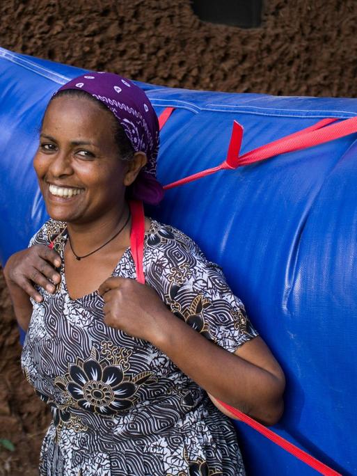 Eine Afrikanerin im geblümten Kleid trägt einen riesigen knallblauen Plastikrucksack voller Biogas auf dem Rücken.