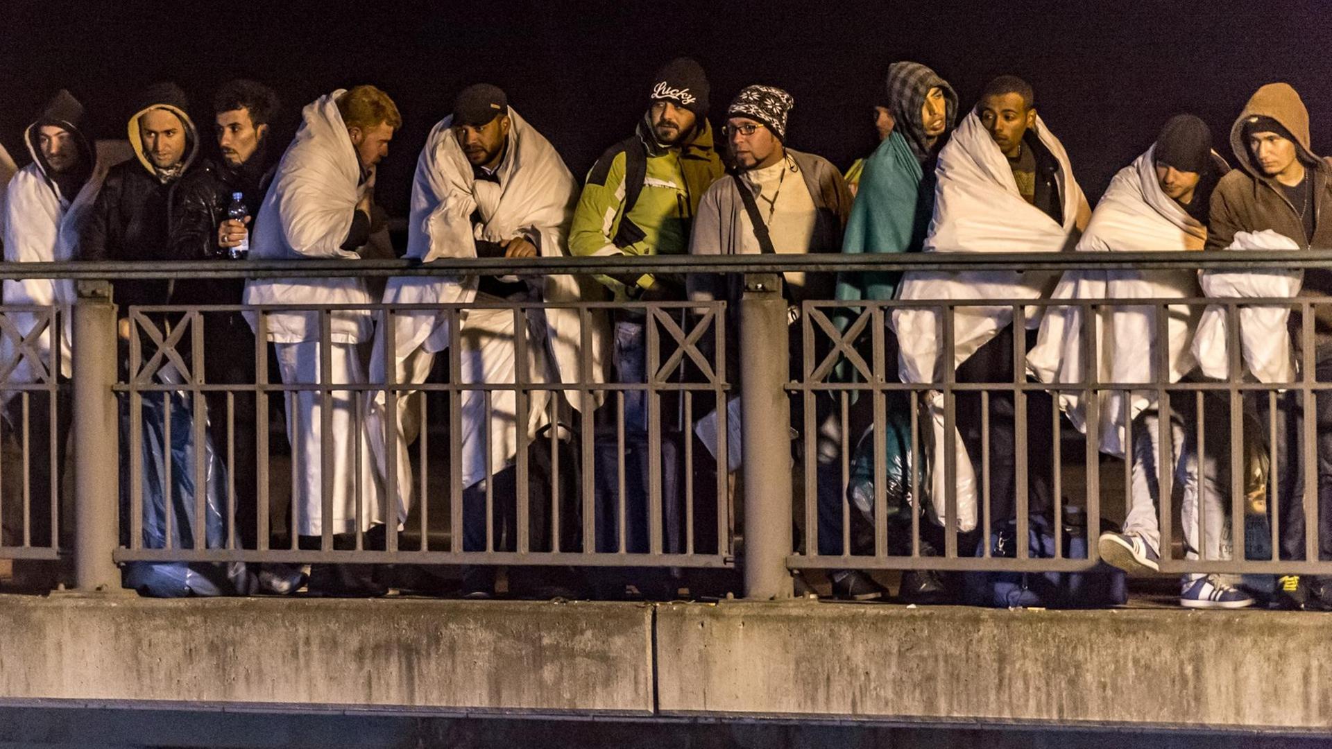 Flüchtlinge stehen nachts in Decken gehüllt auf einer Brücke.