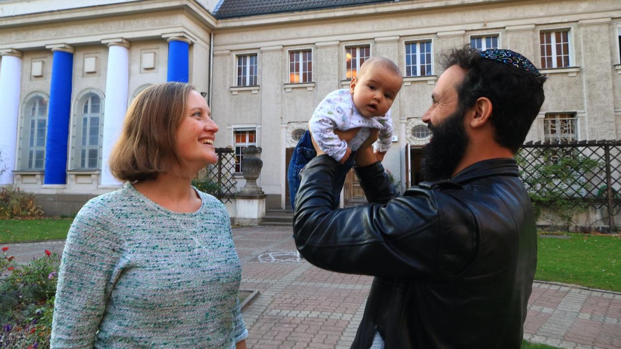 Nina Peretz, blond, im hellblauen Pullover, und ihr Mann Dekel, mit schwarzem Bart in einer schwarzen Lederjacke, stehen einander gegenüber vor einem Portal mit blauen Säulen. Dekel Peretz hebt das Baby in die Höhe.