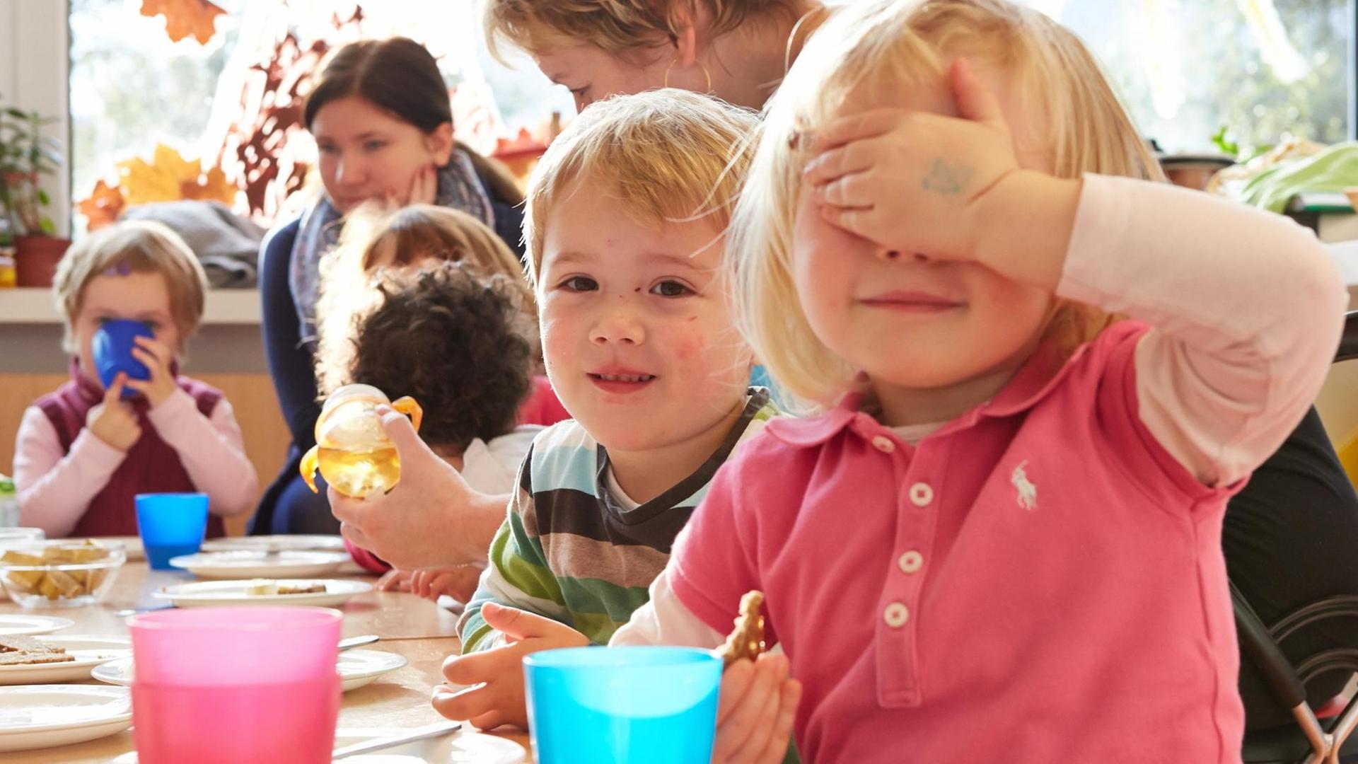 Ein Kind in einer Kita hält sich eine Hand vor die Augen und lacht. Dahinter sitzen weitere Kinder an einem Frühstückstisch.