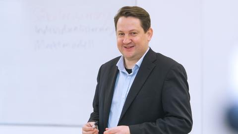 Dr. phil. Alexander Yendell, steht vor einer weißen Wand und lächelt in die Kamera, er hat einen Anzug an und einen Stift in der Hand.
