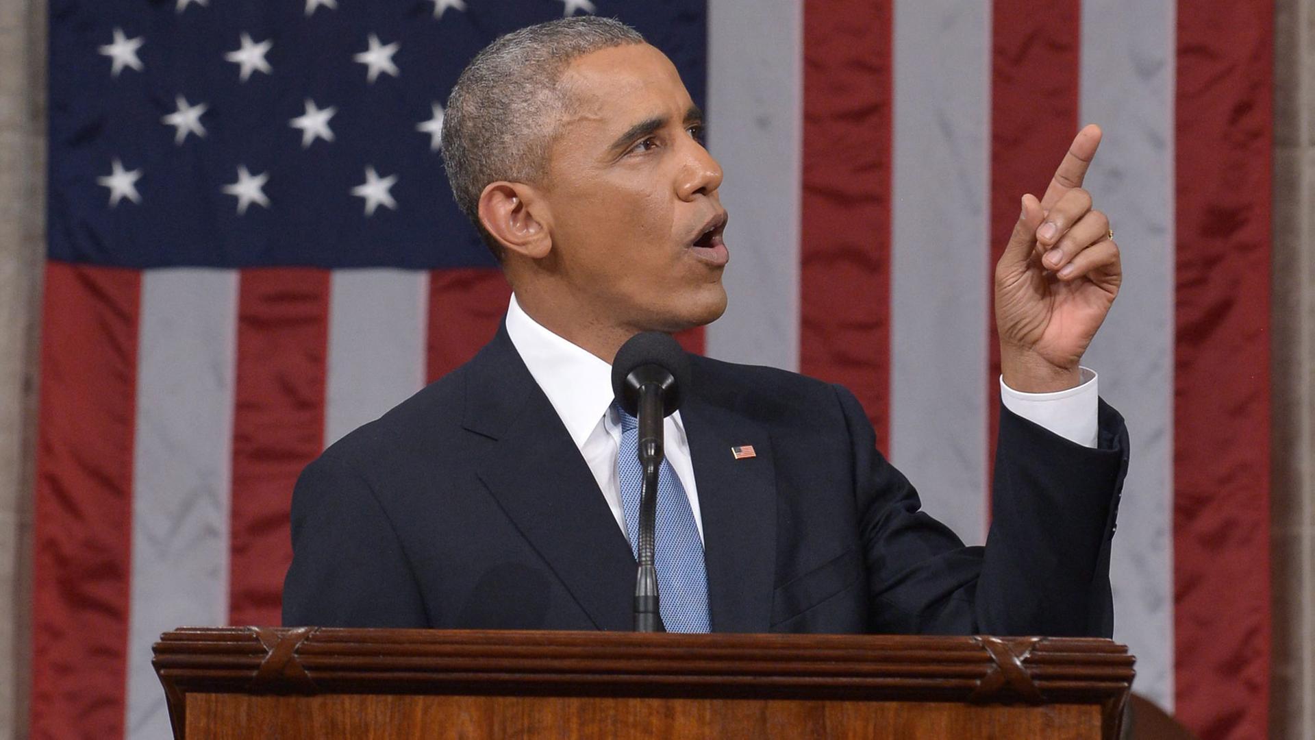 Obama steht vor einer USA-Fahne und zeigt mit dem Finger nach oben.