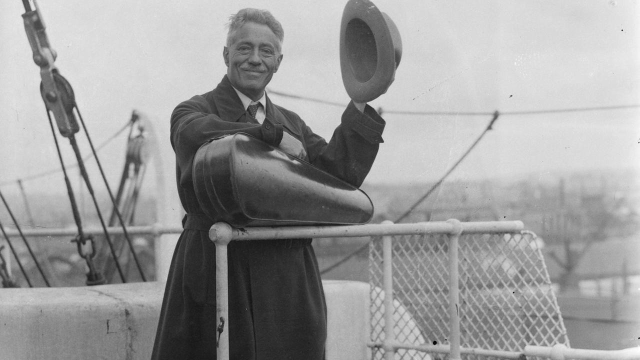 Schwarz-weiß-Foto. Der Geiger Fritz Kreisler 1935 om Southhampton auf der auf dem Weg in die USA. Er steht an der Reling, lüftet seinen Hut, trägt einen dunklen Mantel und hält quer vor sich seinen Geigenkasten.