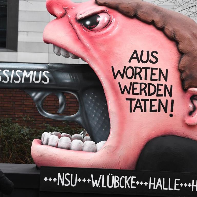 Ein Motivwagen "Rassismus - Aus Worten werden Taten! +++ NSU +++ W. Lübcke +++ Halle +++ Hanau" steht vor dem Rosenmontagszug auf der Straße.
