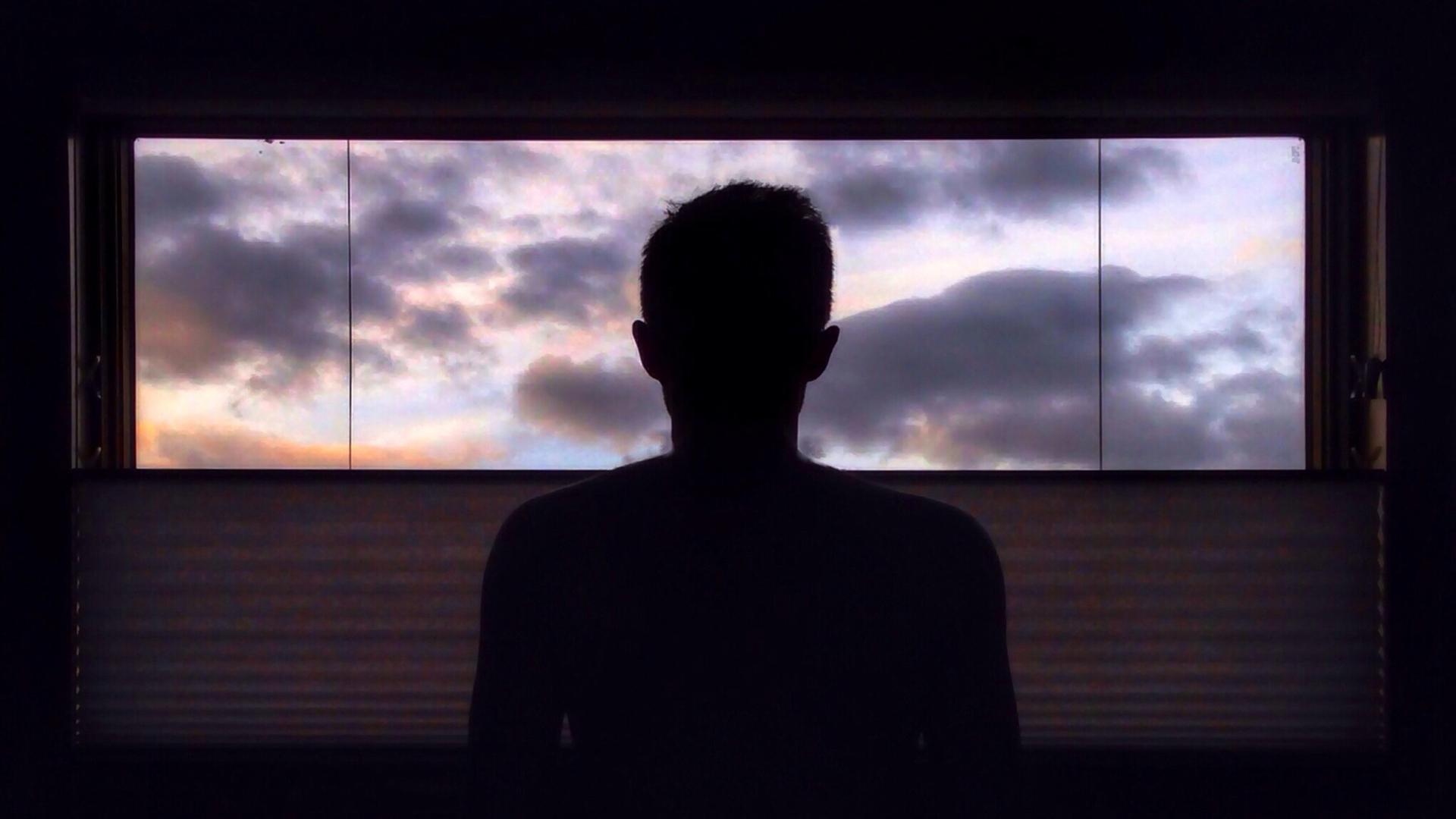 Eine Person sitzt in einem dunklen Raum vor einem Fenster, durch das ein dramatisch bewölkter Himmel zu sehen ist.