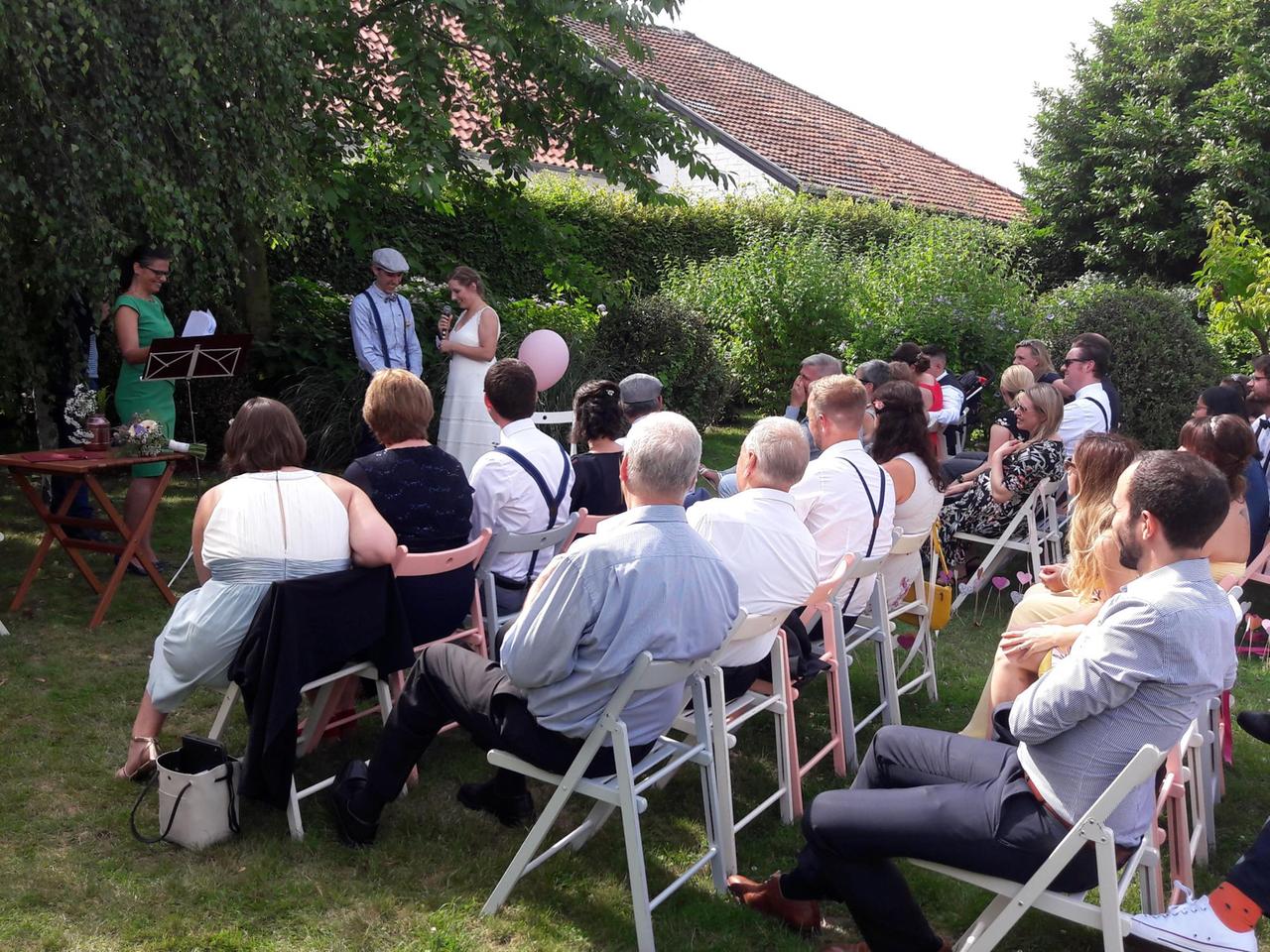 Das Bild zeigt ein Brautpaar, das in einem Garten steht und getraut wird. Im Vordergrund sitzt die Hochzeitsgesellschaft.