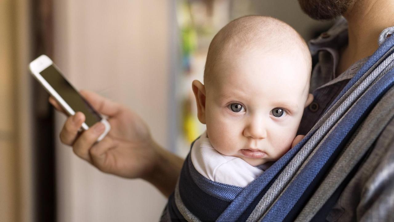 Ein Vater trägt ein Baby an der Brust und schaut dabei auf sein Smartphone