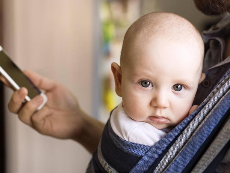Ein Vater trägt ein Baby an der Brust und schaut dabei auf sein Smartphone