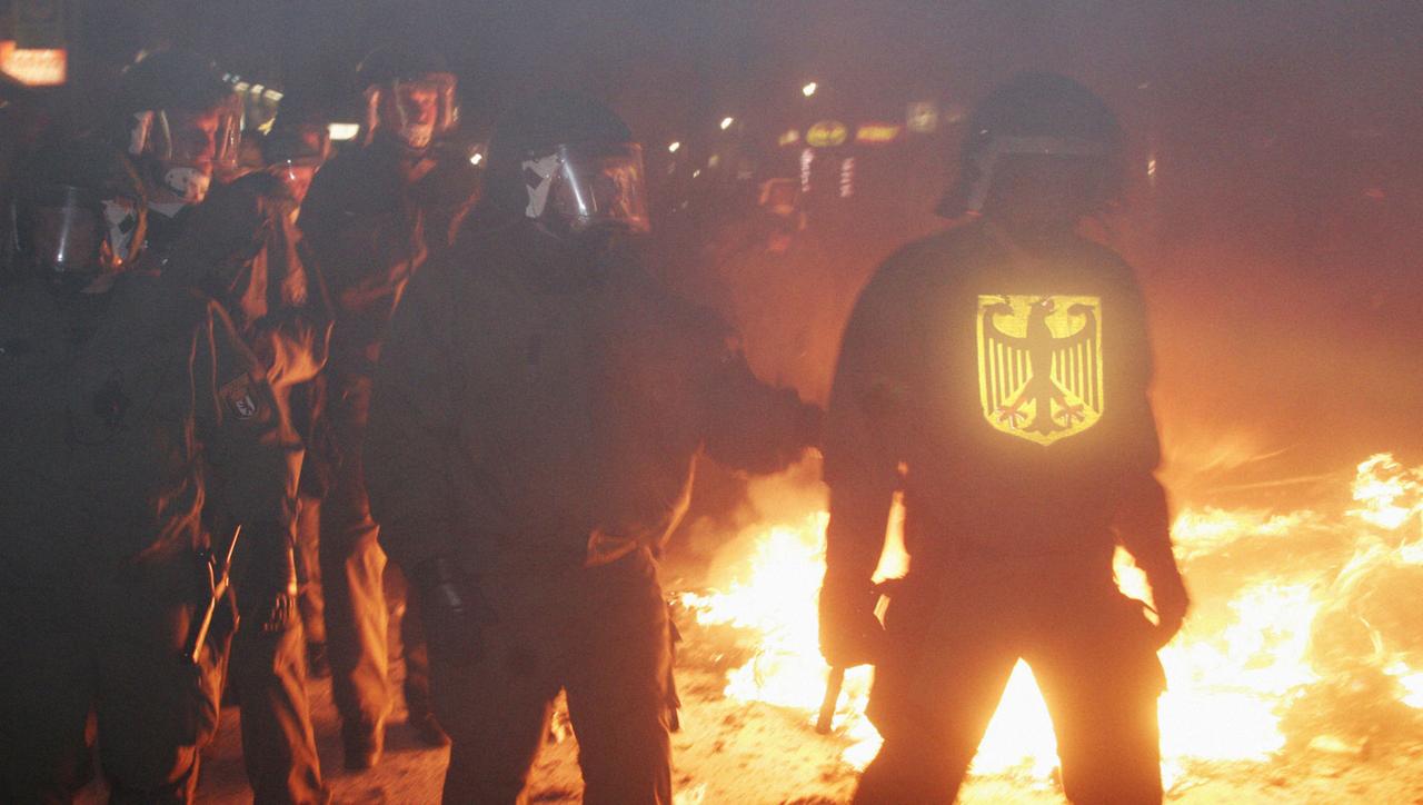 Julius von Bismarck, Fuguration #5 (May Day Riot Police), Polizisten in schwerer Uniform und mit Helmen stehen bei Nacht vor einer am Boden brennenden Flüssigkeit