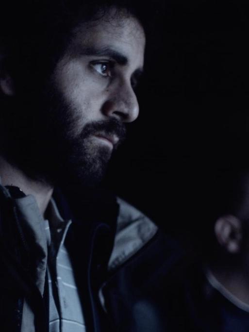 Filmsszene aus dem Kurzfil "Proll!. Murat (Erol Afsin) steht nehen einer weiteren Person im Dunkeln und schaut zur Seite in Richtung des Lichts.
