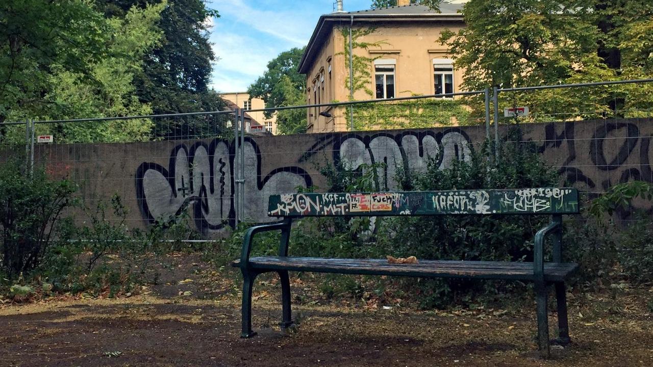 Eine heruntergekommene Parkbank steht vor einem Bauzaun, der eine mit Graffiti besprühte Mauer abschirmt, hinter der ein Gebäude hervorragt. 