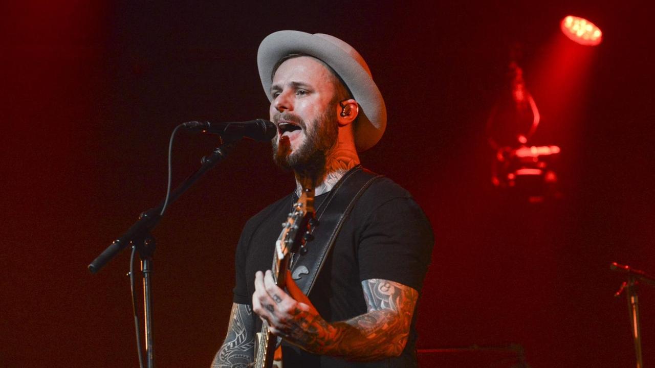 Ein Mann mit Gitarre steht auf einer Bühne, trägt einen Hut und singt in ein Mikrofon. Im Hintergrund leuchtet ein roter Scheinwerfer.
