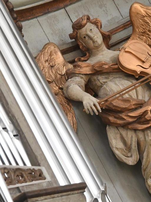 Blick auf eine historische Engelsfigur, die eine goldene Geige spielt und direkt neben den neuen Orgelpfeifen steht.
