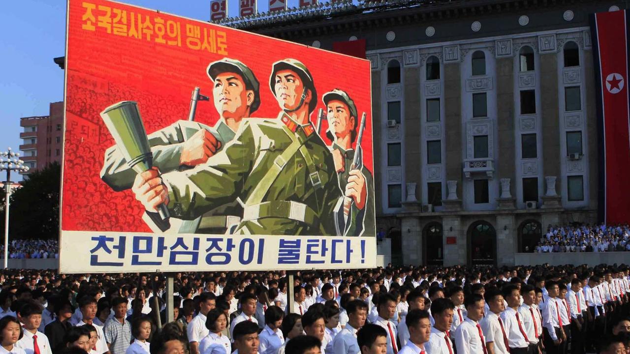 Nordkoreaner in weißen Hemden und roten Krawatten laufen in Reih und Glied über den Platz. Sie tragen ein großes Propaganda-Transparent mit der Abbildung nordkoreanischer Soldaten.