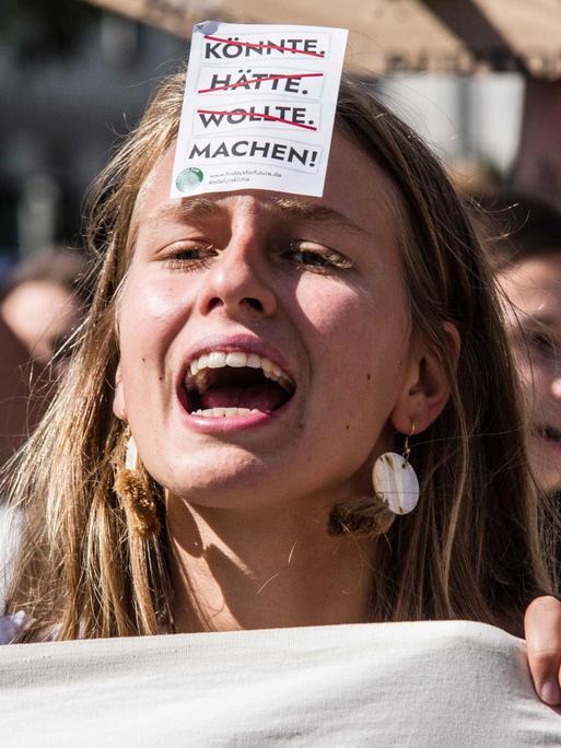 Eine junge Frau mit einem offenen Mund steht inmitten des Demonstrationszuges der fridays for future, einen Flyer auf ihrer Stirn klebend. Die Wörter könnte, hätte, wollte stehen durchgestrichen darauf und das Wort machen mit einem Ausrufezeichen dahinter.