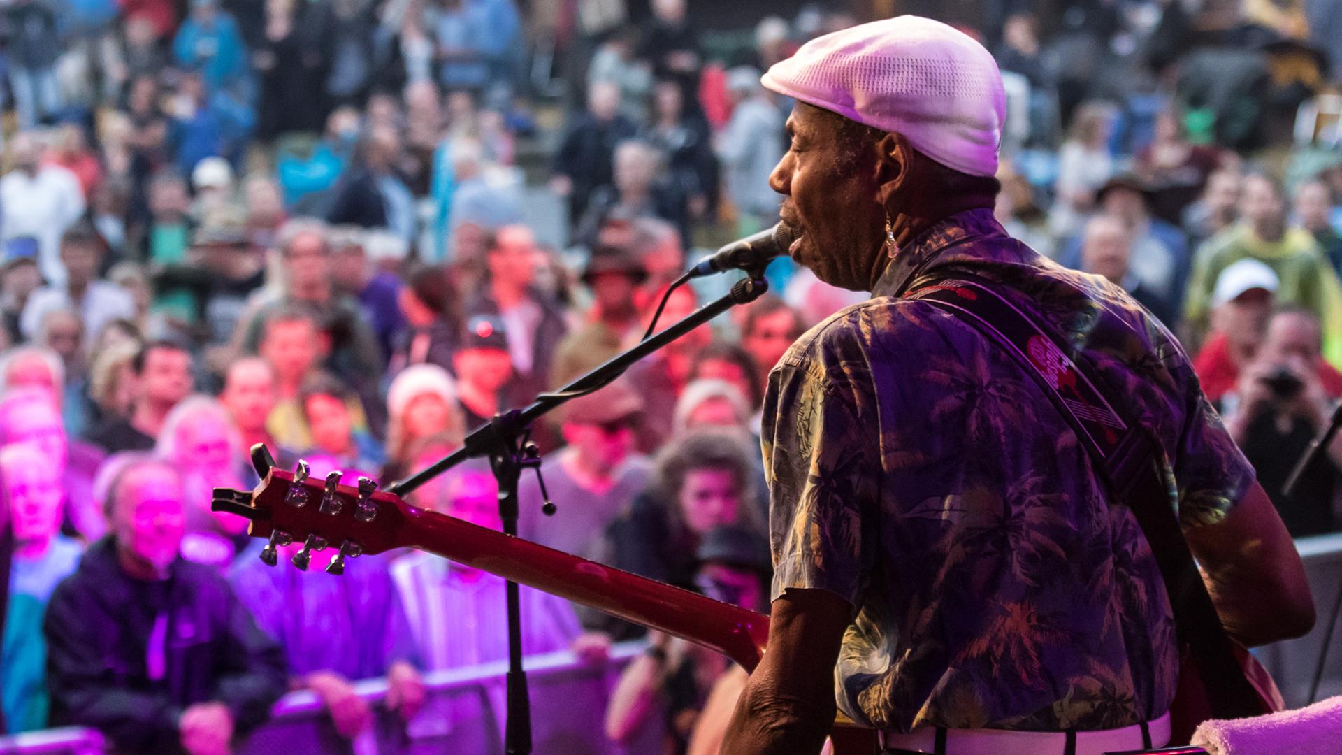 Mann mit Gitarre in der Hand steht mit dem Rücken zur Kamera auf einer Bühne, vor ihm steht Publikum.