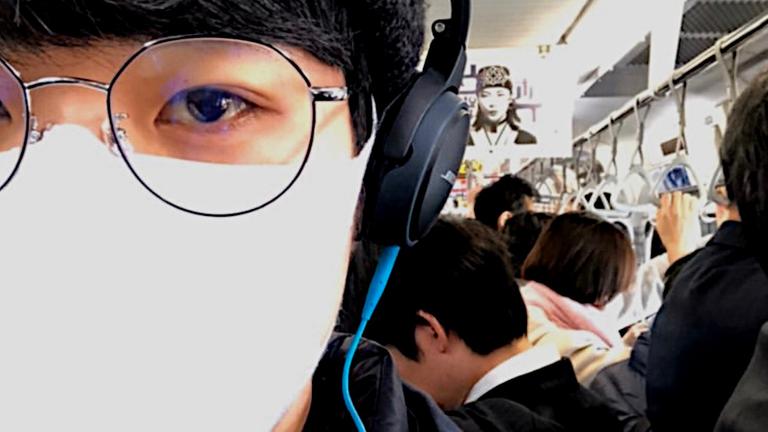 Selfie in einer U-Bahn: Ein junger Mann mit Mundschutz, Kopfhörern und Brille, daneben zahlreiche Fahrgäste