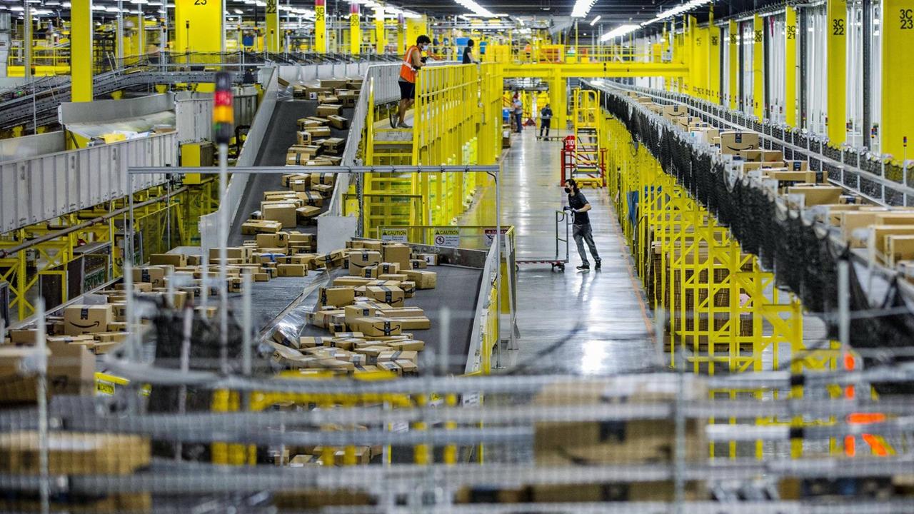 Einblick in ein Amazon-Logistikstandort in den USA. Mitarbeiter tragen Masken, Förderbänder transportieren Pakete unterschiedlicher Größen.