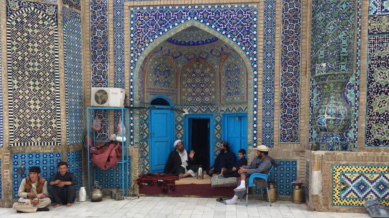 In der Blauen Moschee in Mazar-e Sharif sitzen einige Männer auf Sitzkissen oder dem Boden. Die Wände der Moschee sind mit blauen Kacheln gefliest.