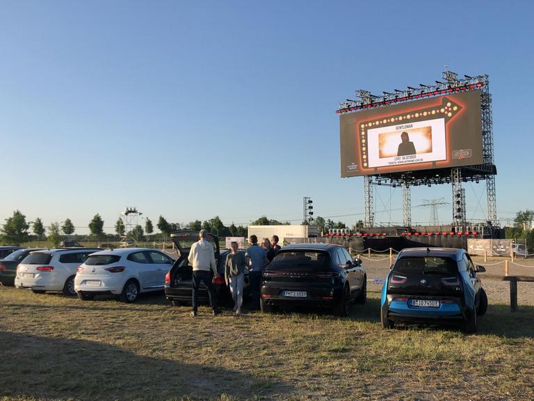 Aufnahme aus einem Autokino, mit Autos im Vordergrund und der Leinwand im Hintergrund, vor blauem Himmel.