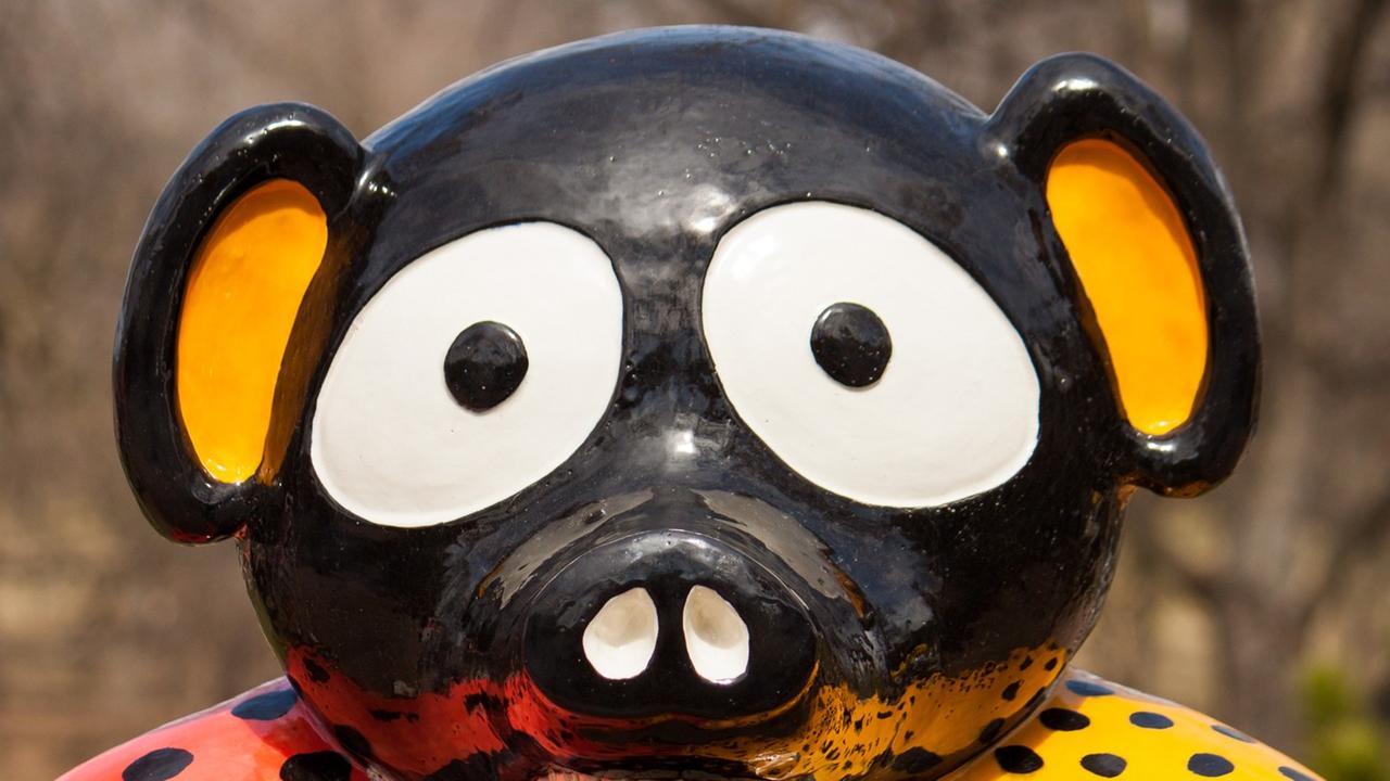 Eine Maske (einem Bär oder Schwein ähnlich) mit schwarzem Gesicht, orangenen Ohren und großen weißen Augen schaut in die Kamera.