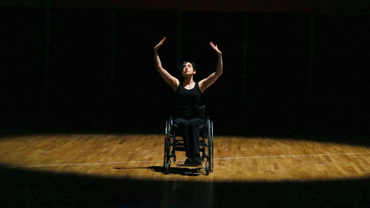 Eine Tänzerin im Rollstuhl performt im Lichtkegel einer sonst dunklen Bühne und streckt ihre Hände in die Höhe.