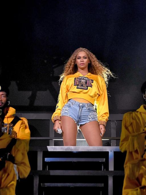 Beyoncé steht in abgerissenen Jeans und einem knallgelben Pullover auf der Bühne und schaut ins Publikum.