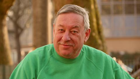 Ein Mann mit kurzen grauen Haaren lächelt verschmitzt in die Kamera. Er trägt einen grünen Pullover, im Hintergrund sind Baumstämme und ein Gebäude im Abendlicht zu sehen.