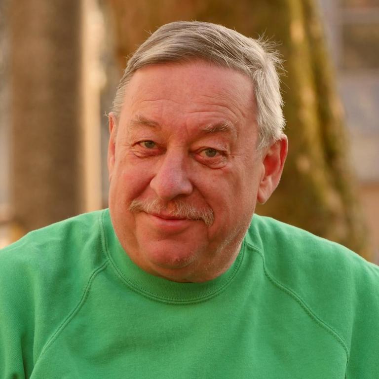 Ein Mann mit kurzen grauen Haaren lächelt verschmitzt in die Kamera. Er trägt einen grünen Pullover, im Hintergrund sind Baumstämme und ein Gebäude im Abendlicht zu sehen.