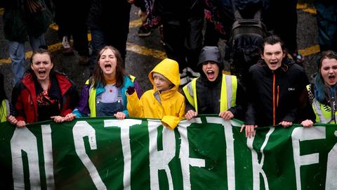 Greta Thunberg hält mit jungen Demonstrierenden ein grünes Banner. Die anderen Jugendlichen rufen etwa. Thunberg schaut zur Seite und winkt.