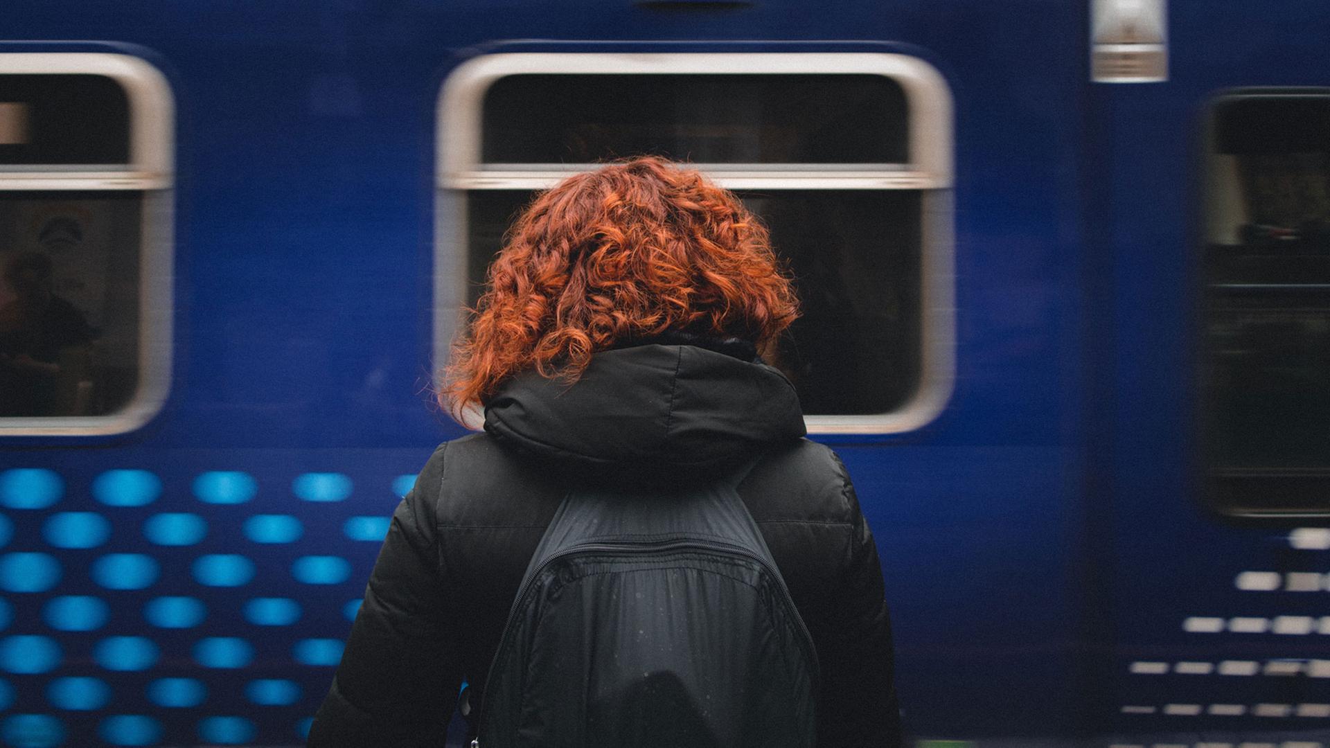 Eine rothaarige Frau steht mit dem Rücken zum Fotografen auf einem Bahnsteig, dahinter ein blauer Zug