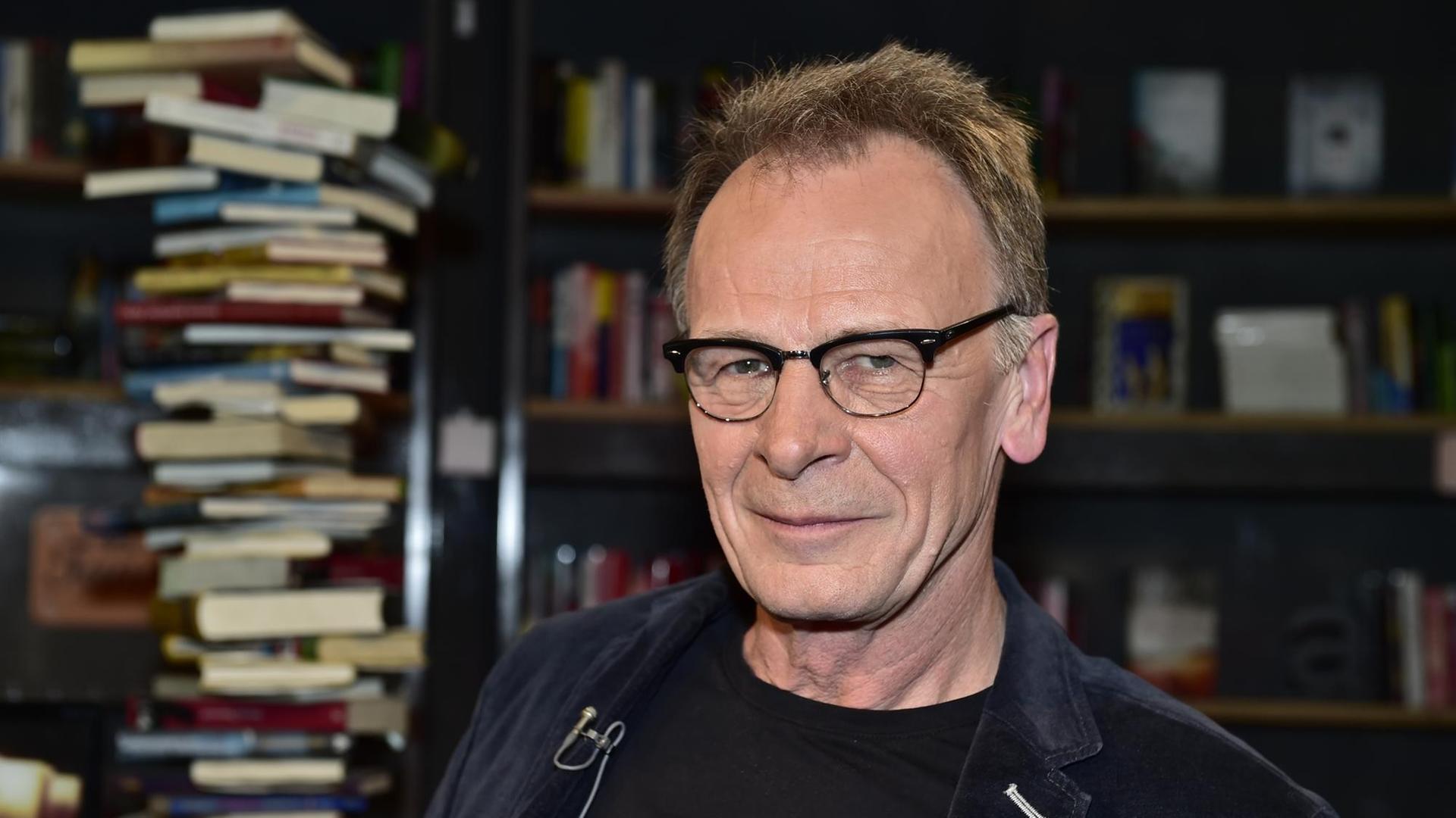 Josef Haslinger abgebildet vor übereinander gestapelten Büchern und einem vollen Bücherregal. Er trägt eine Brille.