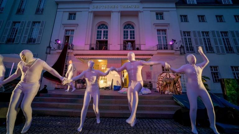 Vier in silberfarbene Latex-Anzüge gekleidete Menschen rennen die Treppe vom Deutschen Theater herunter und halten sich dabei an den Händen.