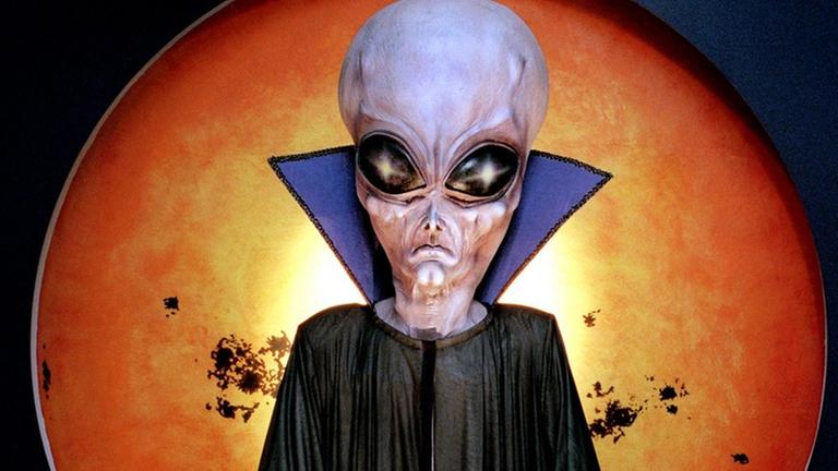 Die Figur eines Außerirdischen mit hoher Stirn und großen Augen vor einer Planeten-Abbildung
