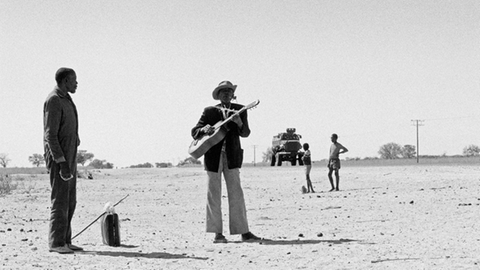 Schwarz-Weiß-Aufnahme zweier Männer auf sandigem Boden. Der eine spielt Gitarre.