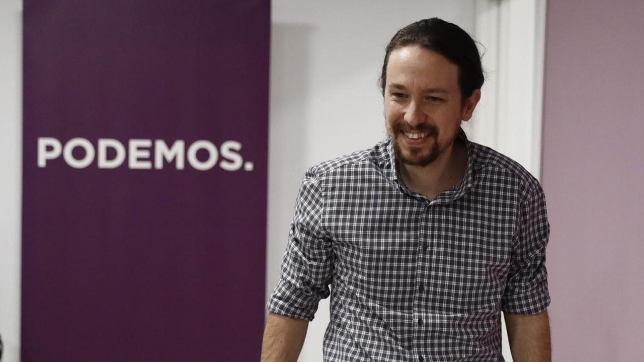 Ein junger Mann mit kurzem Bart, dunklem Zopf und kariertem Hemd steht lächelnd vor dem "Podemos"-Logo.