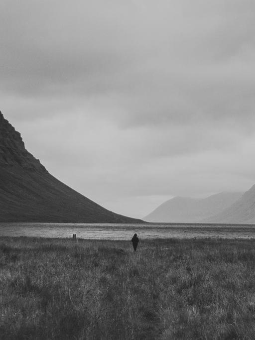 SW-Foto einer isländischen Landschaft mit Berg und See, nahe des Wasserfalls Dynjandi. In der Bildmitte ist die Silhouette eines Menschen zu sehen.