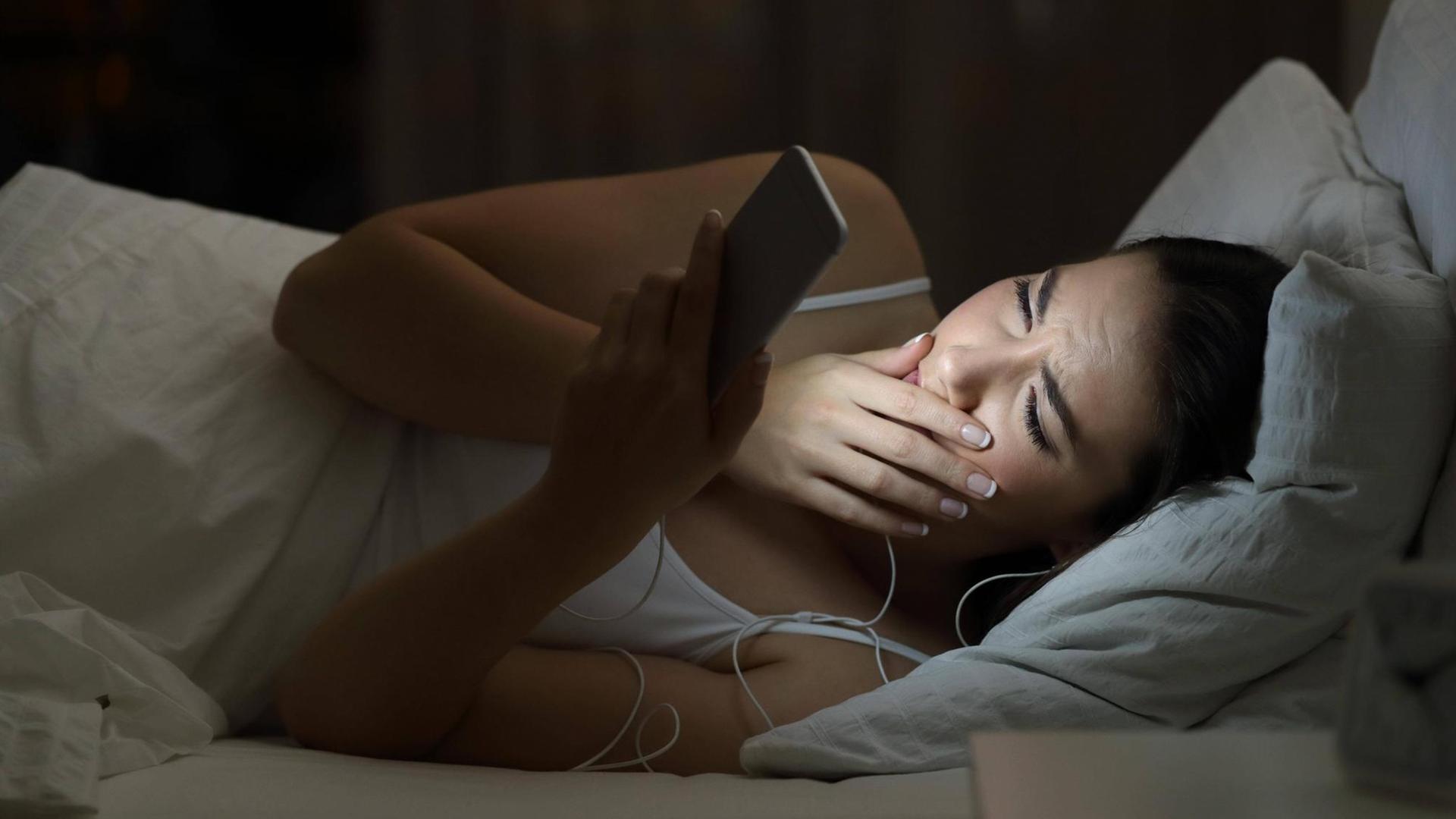 Eine junge Frau liegt im Bett, gähnt und schaut auf ihr Handy.