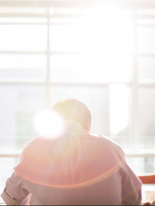 Ein Mann sitzt an einem Konferenztisch mit dem Gesicht zur Fensterfront, durch die die Sonne scheint.