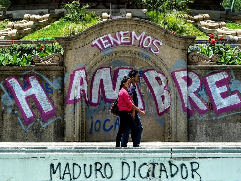 Ein Paar läuft auf der Straße. Dahinter zu sehen: Graffiti mit dem Text "Wir haben Hunger" und "Diktator Maduro" in Venezuelas Hauptstadt Caracas.