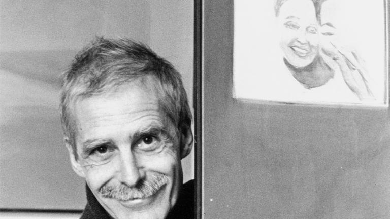 Zu sehen ist eine Schwarz-weiß-Fotografie von Hannes Binder. Er steht in einem Türrahmen und lächelt frontal in die Kamera.