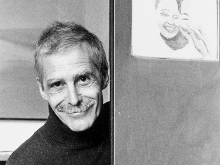 Zu sehen ist eine Schwarz-weiß-Fotografie von Hannes Binder. Er steht in einem Türrahmen und lächelt frontal in die Kamera.