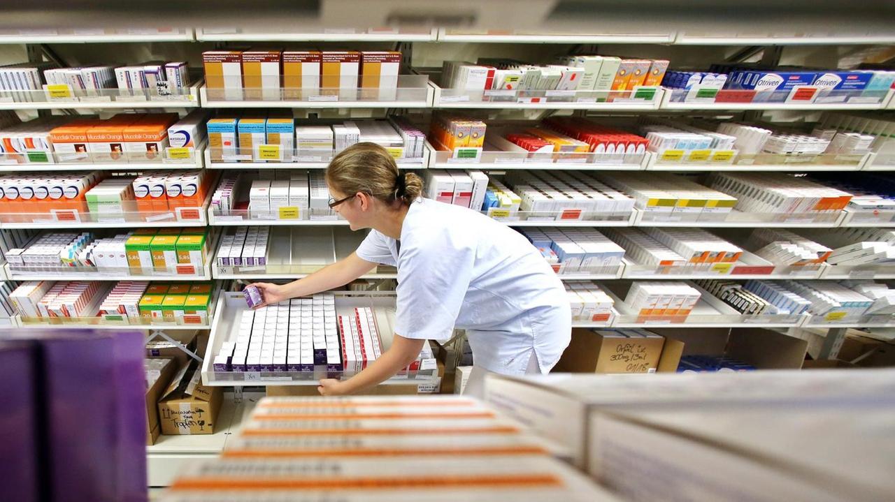 Die Mitarbeiterin einer Apotheke steht vor Regalen mit Medikamenten und nimmt ein Präparat aus einer Schublade.