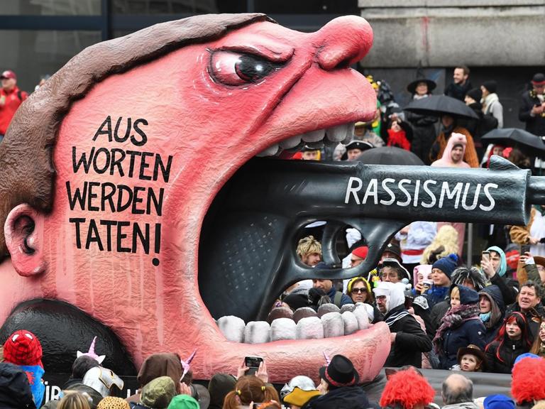Zu sehen ist ein Motivwagen auf dem Karnevalsumzug in Düsseldorf, der einen aufgerissenen Mund zeigt, aus dem dem ein Pistolenlauf ragt. Auf dem Wagen steht der Schriftzug "Rassismus - Aus Worten werden Taten!".
