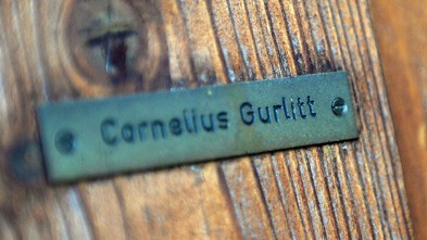 Ein grünes Namensschild auf dem mit schwarzen Buchstaben Cornelius Gurlitt zu lesen ist. Das Schild ist an einen braunen Holzhintergrund (vermutlich eine Tür) geschraubt.